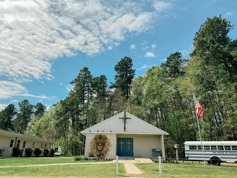 White chapel with a lion mural, a cross on its gable, an American flag and a bus on the House of Prayer grounds