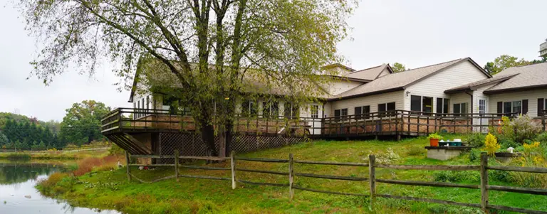 White two-story building with a wooden deck beside a pond and grassy lawn