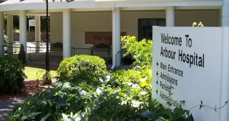 Arbour Hospital entrance with a 'Welcome to Arbour Hospital' sign, shrubs, and a covered walkway