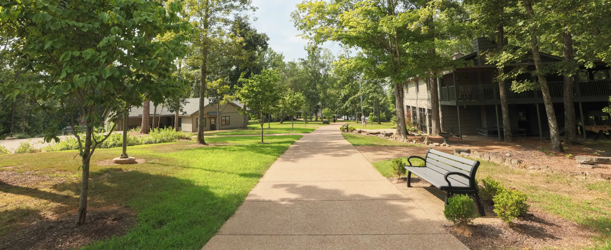 Paved walkway lined with trees and grass leading past wooded cabins, with a bench on the right