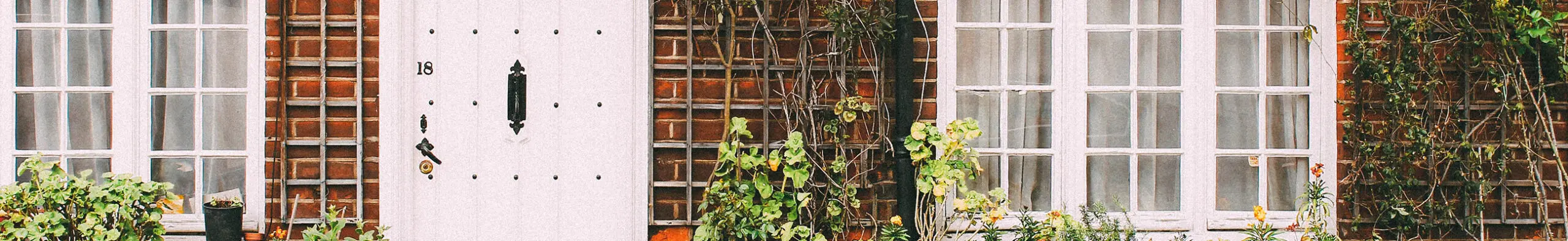 White front door with a house number 18 beside brick walls, windows and climbing plants