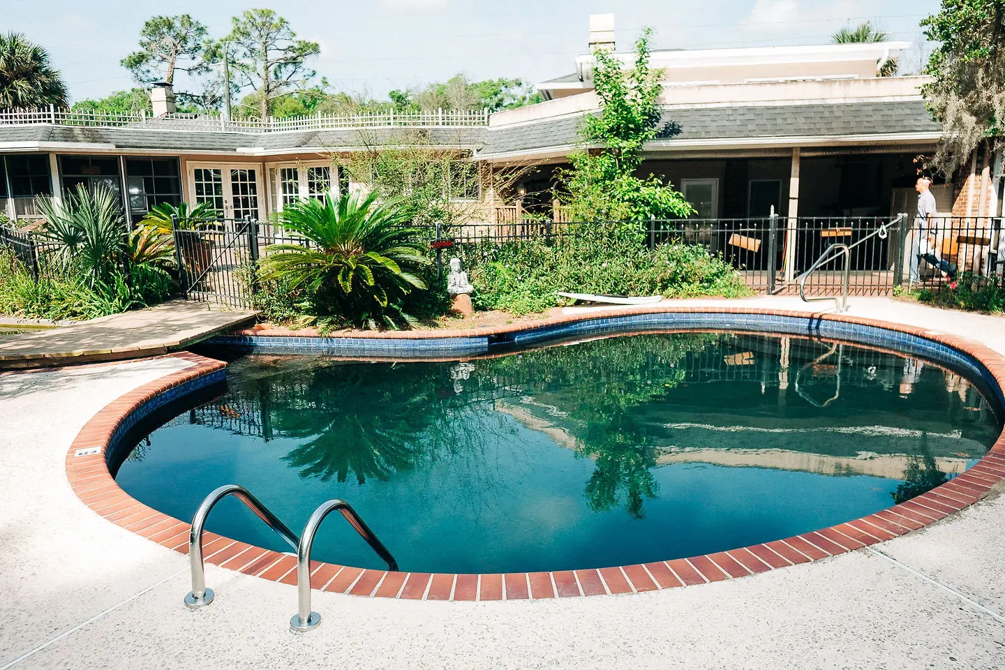 Outdoor pool with tiled edge in front of a single-story building with a covered porch and palm plants