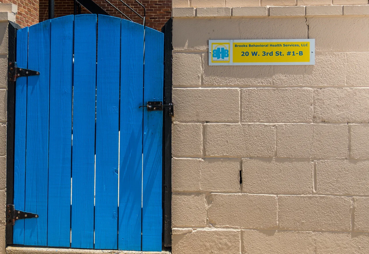 Blue door beside a wall sign reading Brooks Behavioral Health Services, LLC, 20 W. 3rd St. #1-B