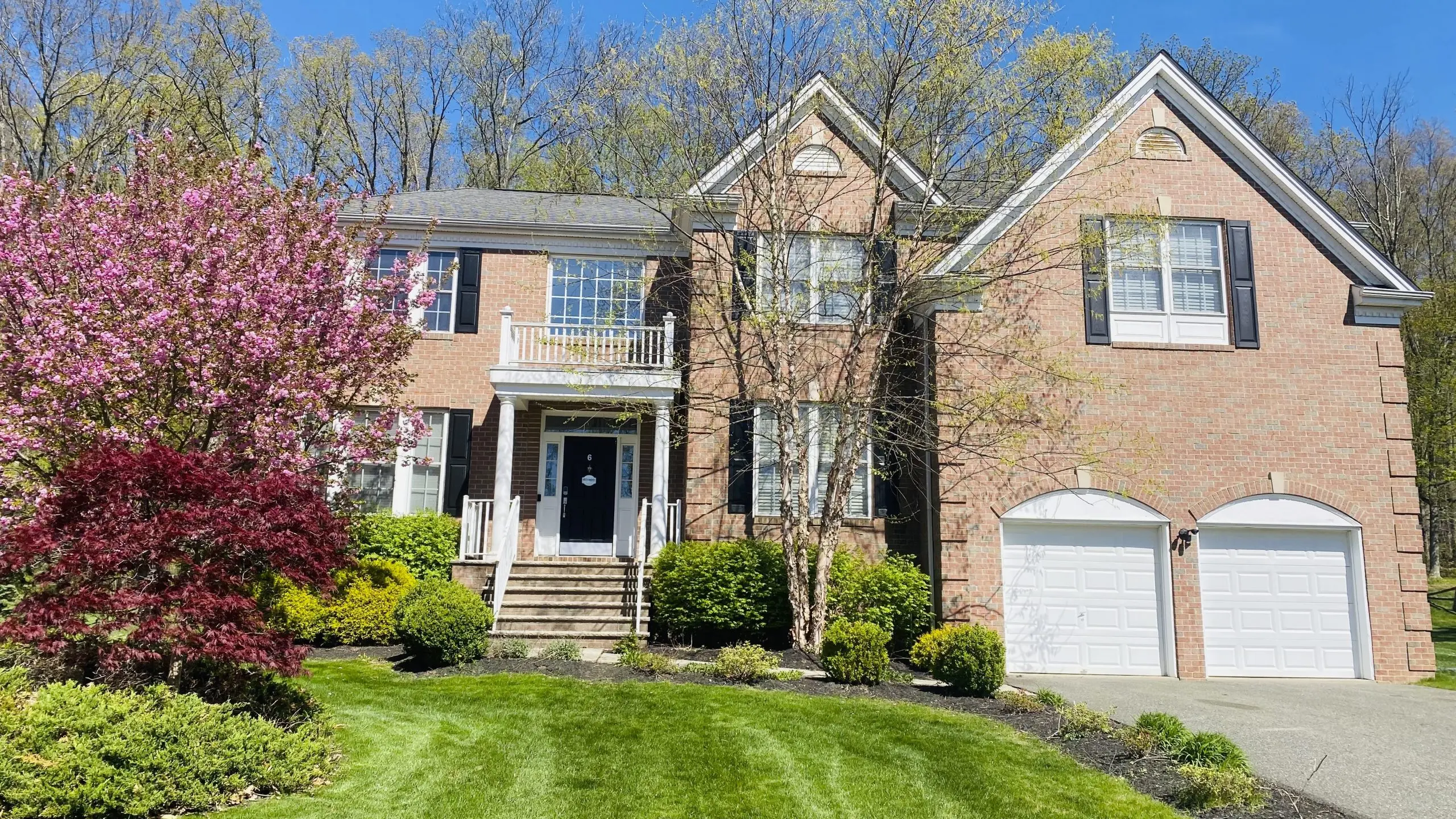 Two-story brick house with a front porch, dark door, and attached two-car garage, surrounded by spring landscaping