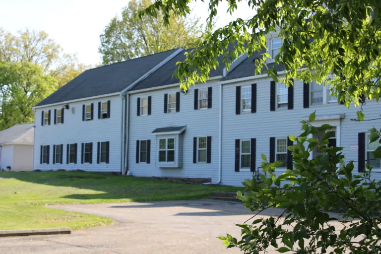 Two-story white building with black shutters, green lawn, and a paved driveway under trees