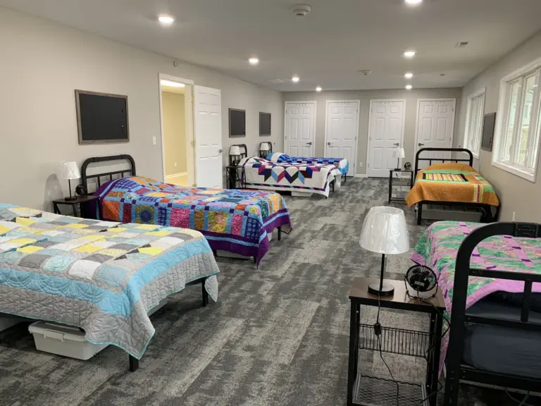 Bedroom with several beds covered in colorful patchwork quilts, lamps, and gray carpet