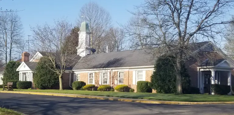 Single-story brick building with a white cupola and columned entrance portico, surrounded by trees and lawn