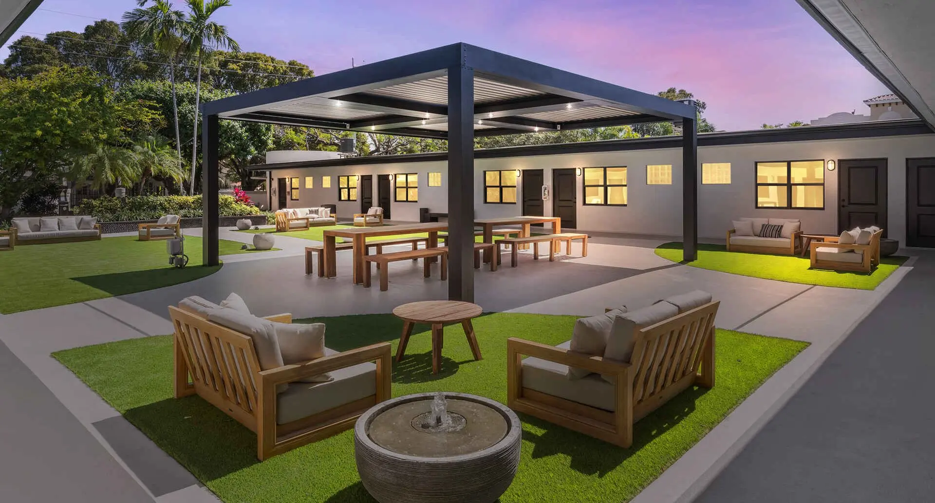 Covered outdoor patio with teak lounge chairs, picnic tables and a stone fountain on artificial grass at dusk