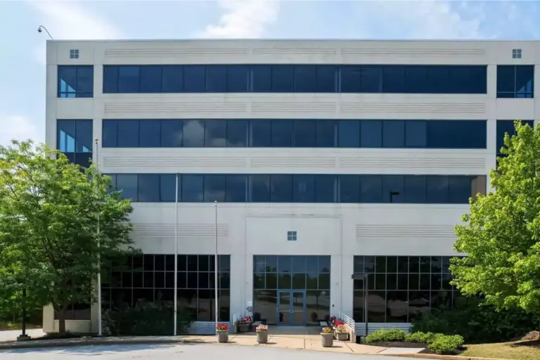 Four-story white office-style building with dark blue windows, central glass entrance, and planters out front