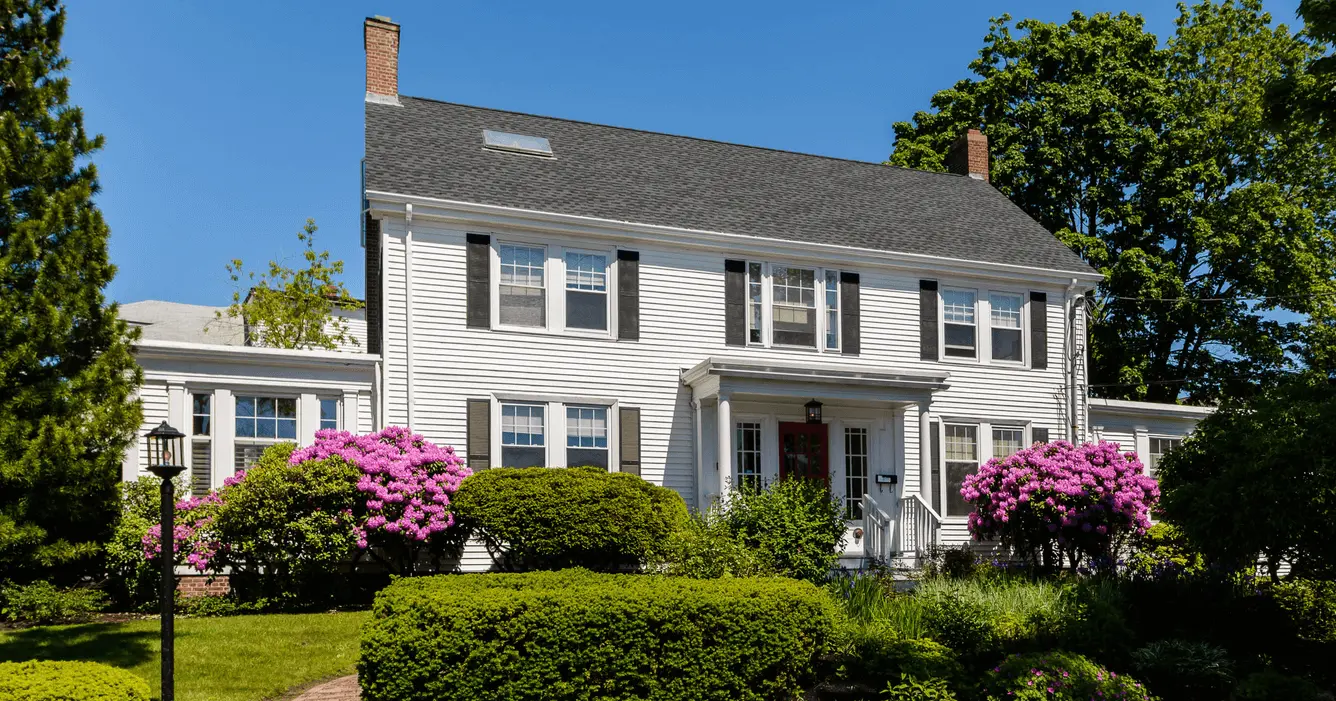White colonial-style house with a gray roof, pink rhododendrons, and a front porch