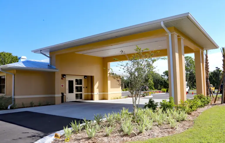Yellow stucco building with a covered entrance drive and landscaped front beds under a clear blue sky