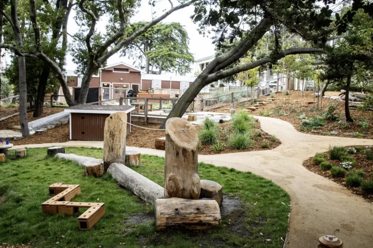 Outdoor play garden with log seats, a wooden play structure, and a brown building in the background