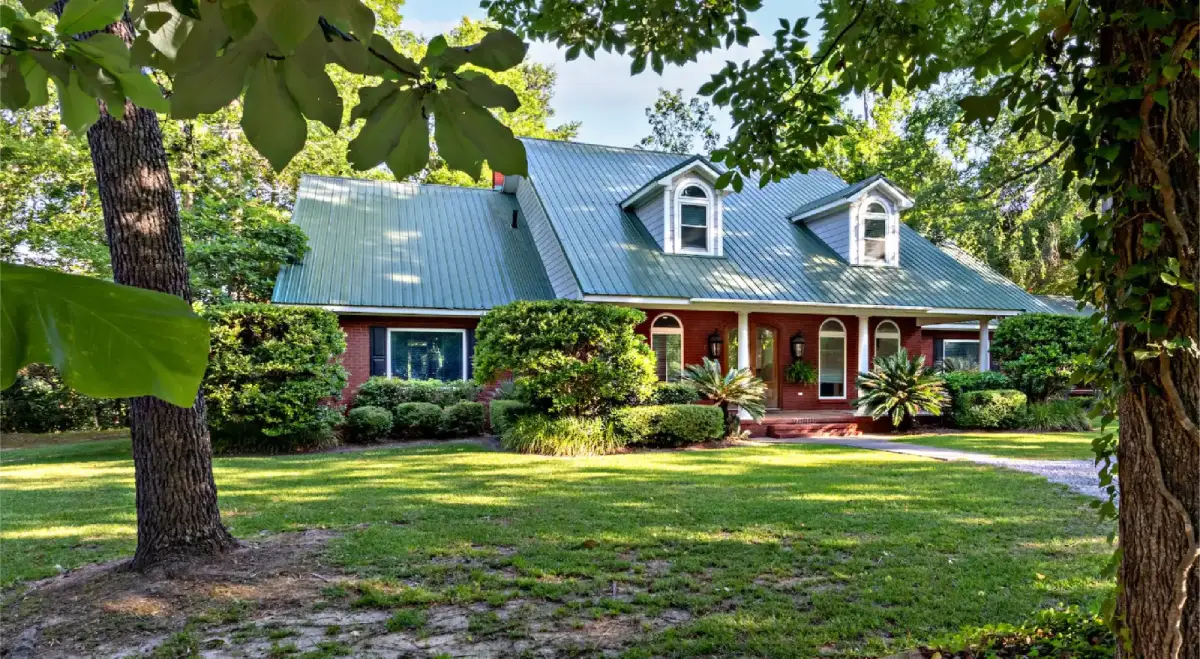Brick house with green metal roof, front porch and lawn surrounded by trees