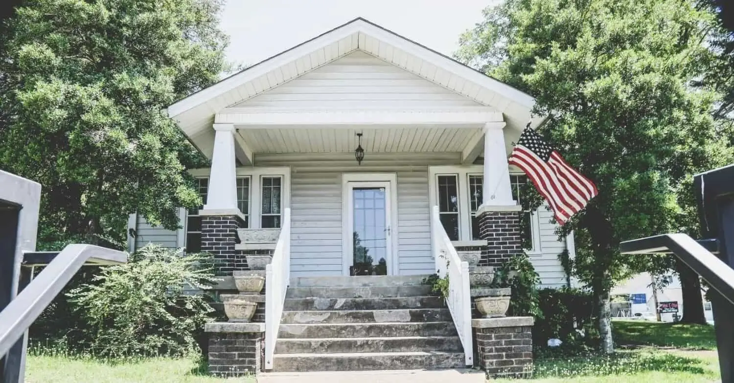 Small white bungalow with a covered front porch, brick columns, and an American flag