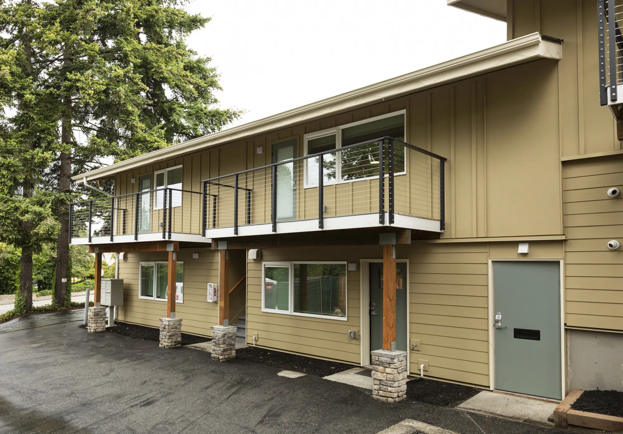 Two-story tan building with balconies, stone piers, and a gray entrance door beside a parking lot