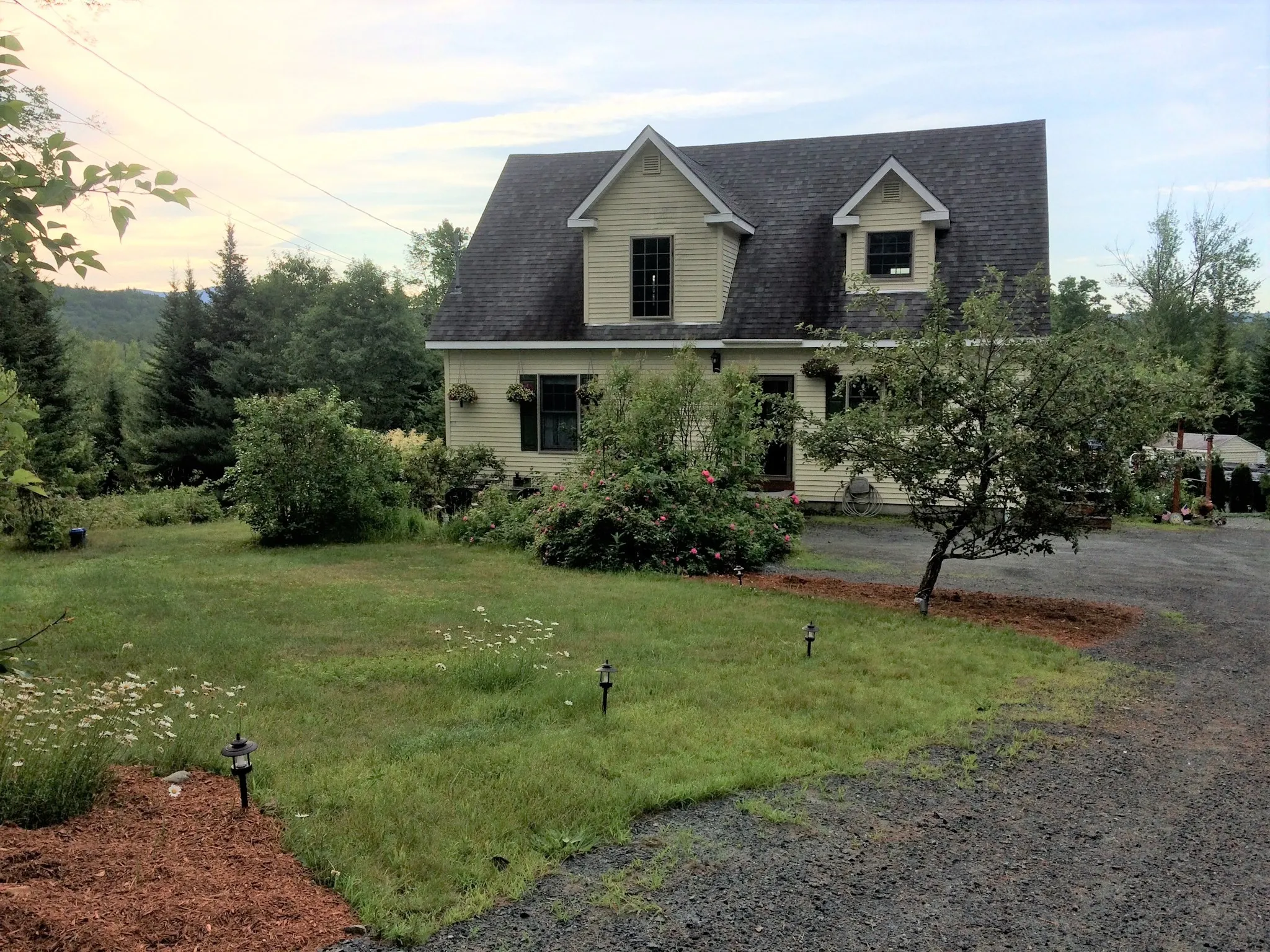 Pale yellow two-story house with dormer windows, a gravel driveway and a landscaped front lawn