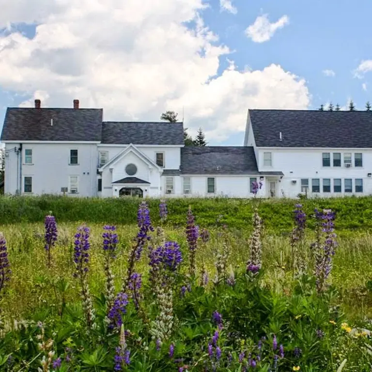 White two-story building with purple lupine wildflowers in the foreground under a partly cloudy sky