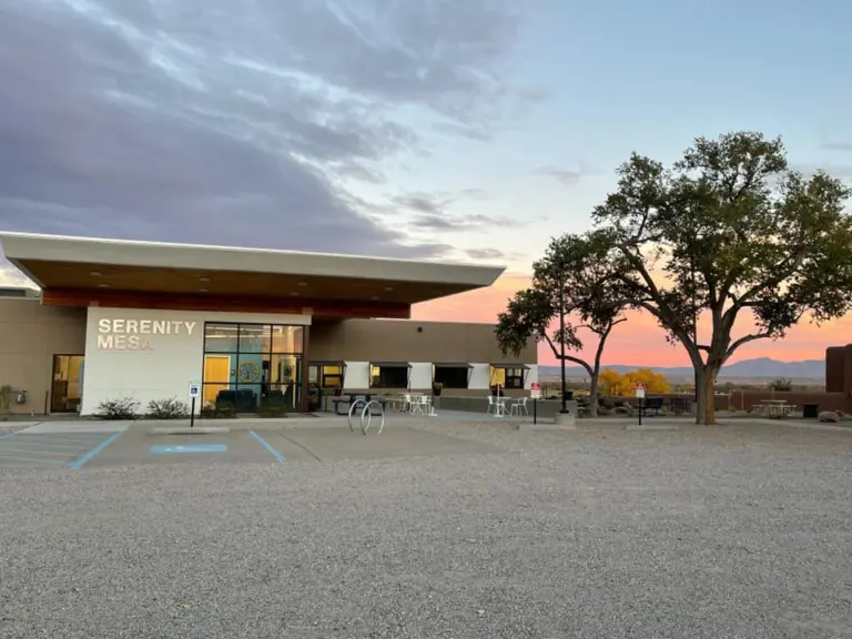 Flat-roofed building with a Serenity Mesa sign at dusk, with trees and a gravel parking area