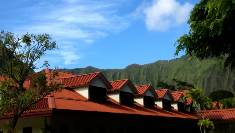Long building with red metal roofs and dormer windows, green mountains and blue sky behind it
