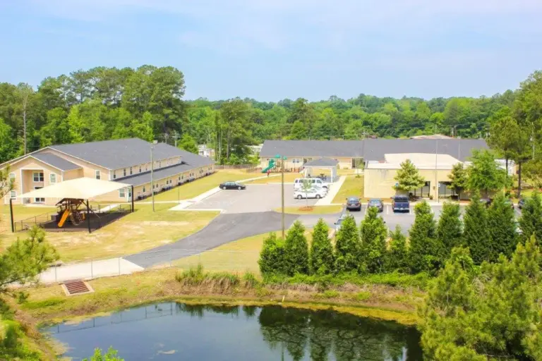 Aerial view of a campus with tan two-story residential buildings, parking lots, trees and a pond