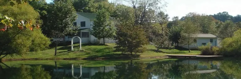 Two-story white building and smaller annex beside a pond surrounded by trees and grass