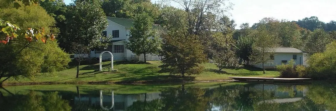 Two-story white building and smaller annex beside a pond surrounded by trees and grass
