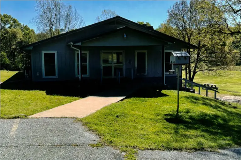 Single-story gray building with a covered entrance, a concrete walkway, and a lawn with a picnic table