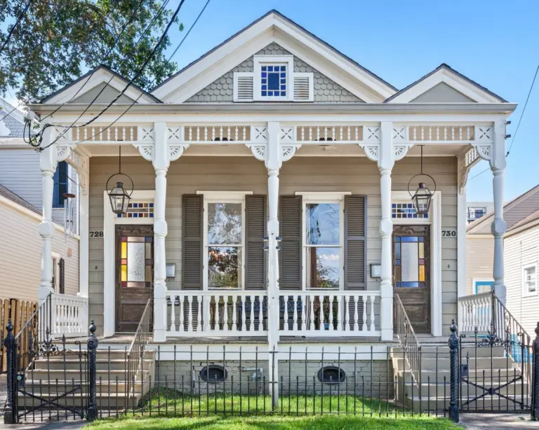 Two-unit white Creole cottage with a front porch, wrought-iron fence, and shuttered windows under a clear blue sky