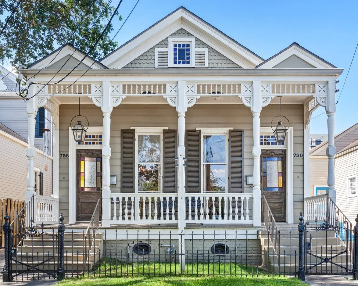 Two-unit white Creole cottage with a front porch, wrought-iron fence, and shuttered windows under a clear blue sky