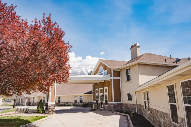 Two-story tan and stone building with a covered entrance drive, autumn tree, and mountains behind