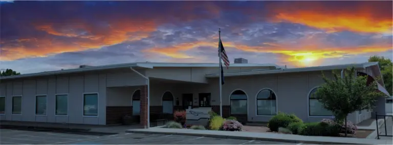 Gray one-story Lifeways building with a flagpole and entrance sign under a sunset sky