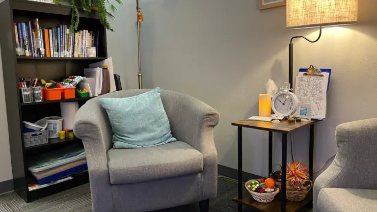 Counseling room with gray armchair, bookshelf, lamp, and side table holding toys and a clock