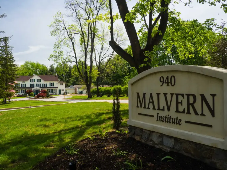 White colonial-style building beside a stone sign reading 940 Malvern Institute, with a lawn and trees