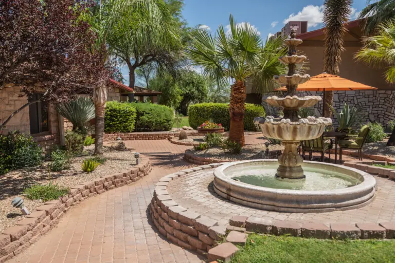 Courtyard with a tiered stone fountain, brick pathways, palm trees, and patio seating beside a stone building