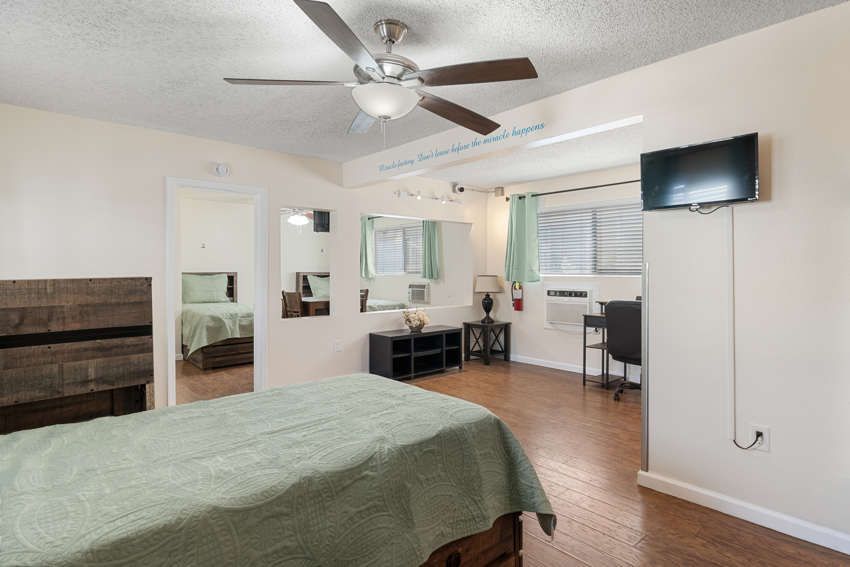 Bedroom with a green quilted bedspread, wall-mounted TV, desk, and a motivational wall quote
