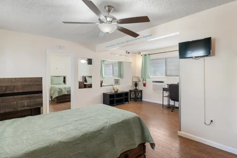 Bedroom with green bedspread, wall-mounted TV and desk, with a wall quote about miracles