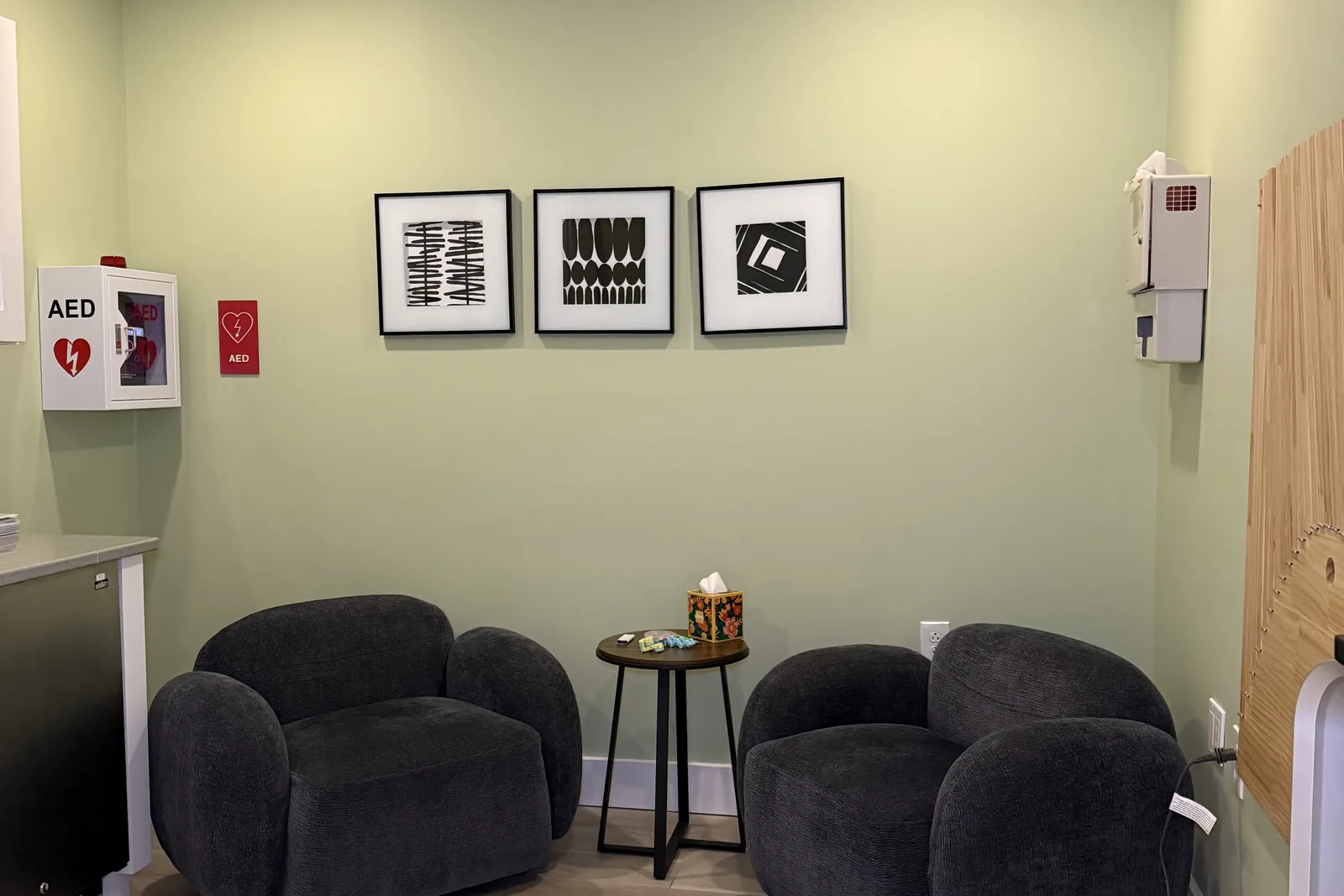 Waiting area with grey lounge chairs, a side table with tissues, framed prints, and an AED cabinet on green walls