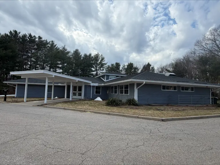 Single-story blue-gray building with a covered entrance and pine trees behind it