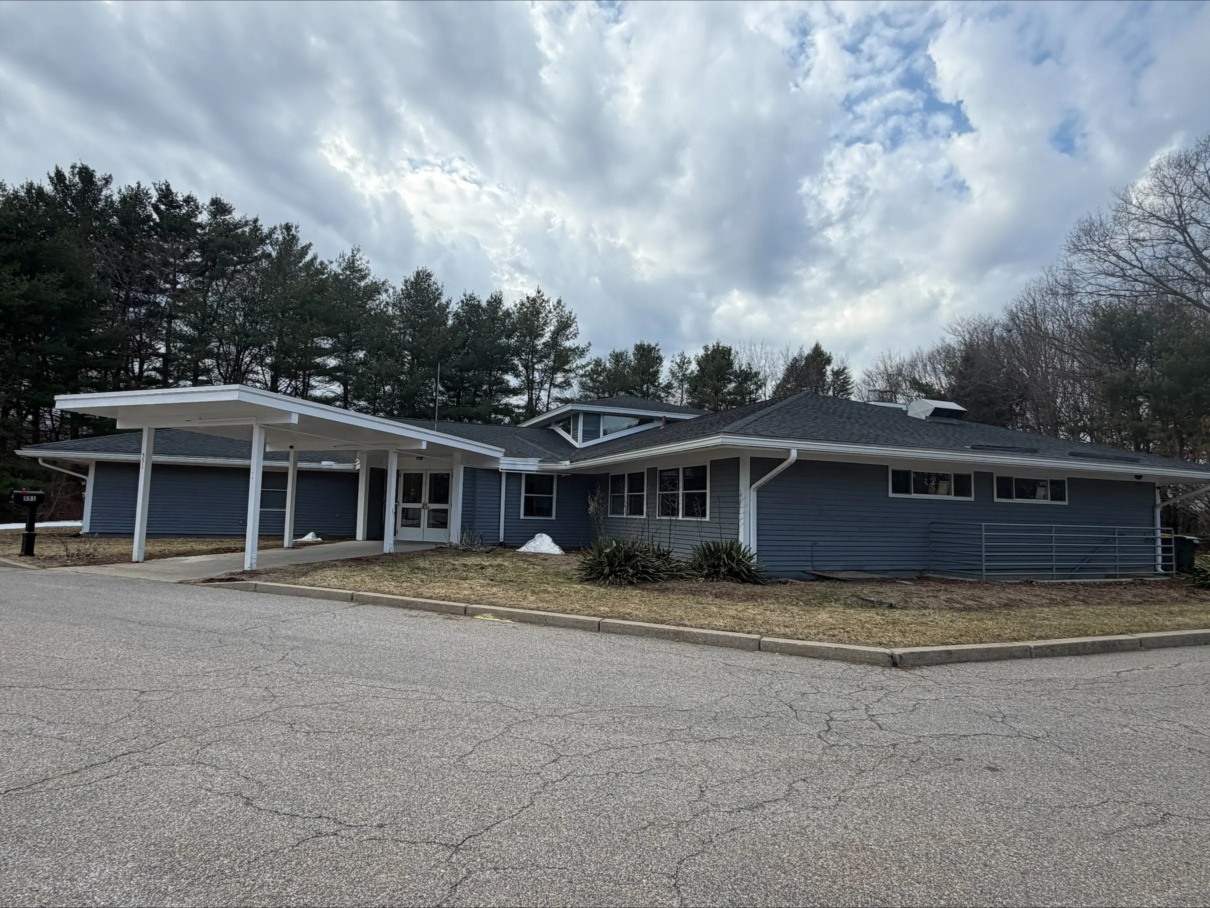 Single-story blue-gray building with a covered entrance and pine trees behind it