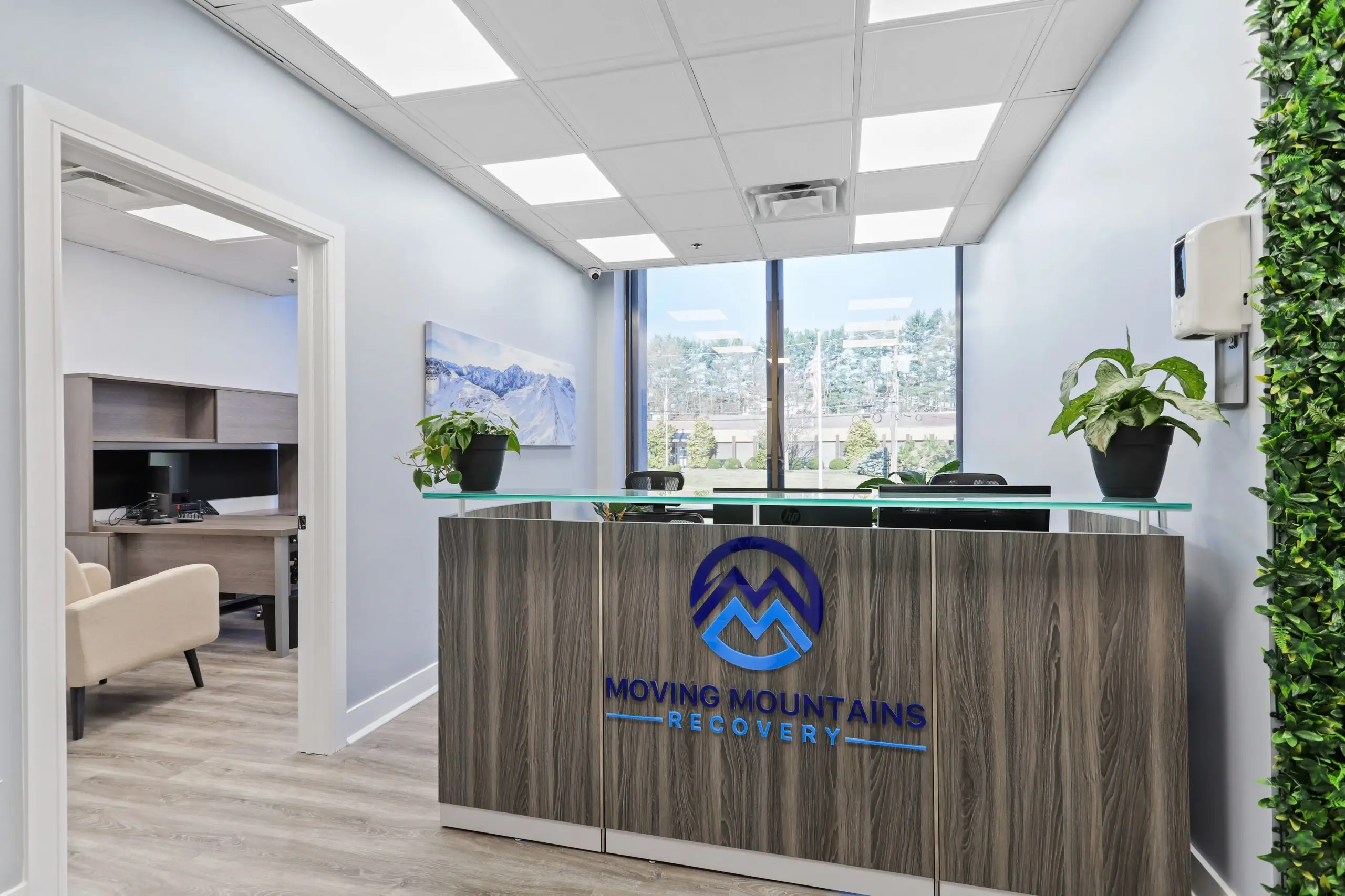 Lobby with Moving Mountains Recovery logo on a wood reception desk, plants, and a window view