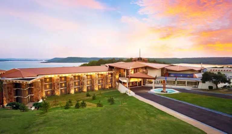 Multi-story campus building with stone facade and fountain beside a lake at sunset
