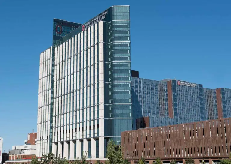 Tall glass and white-panel hospital tower with an Ohio State University Wexner Medical Center sign under a blue sky