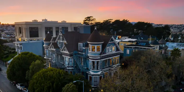 Blue Victorian mansion with a turret at sunset, viewed from above with trees in the foreground