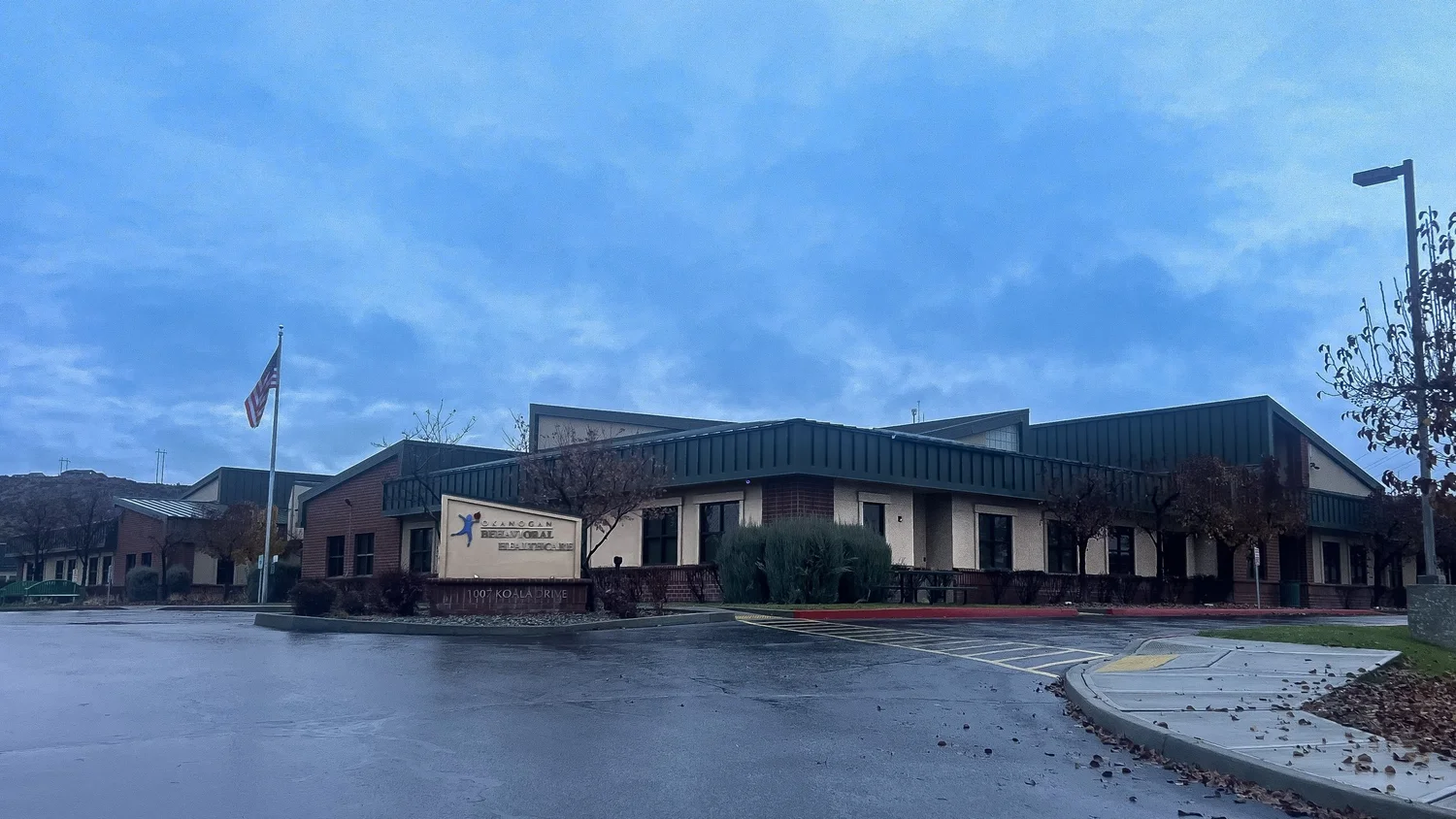 Single-story building with a sign reading Okanogan Behavioral Healthcare under a cloudy blue sky