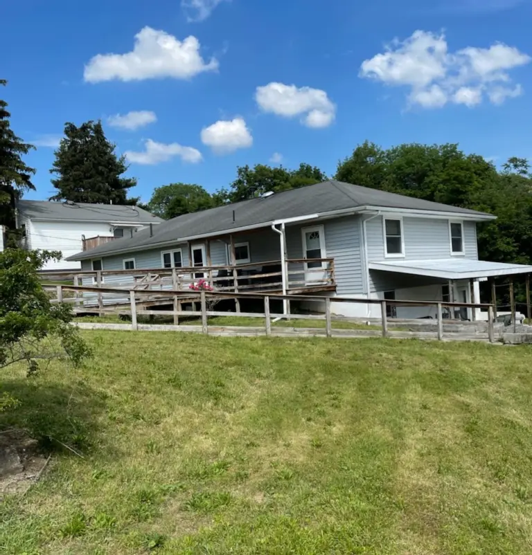 Gray two-story house with a wooden deck and ramp on a grassy hillside under a blue sky