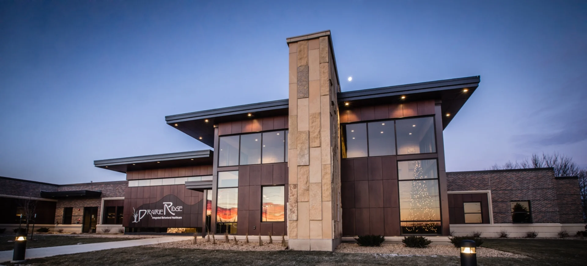Modern two-story building with a stone tower and Prairie Ridge Integrated Behavioral Healthcare sign at dusk