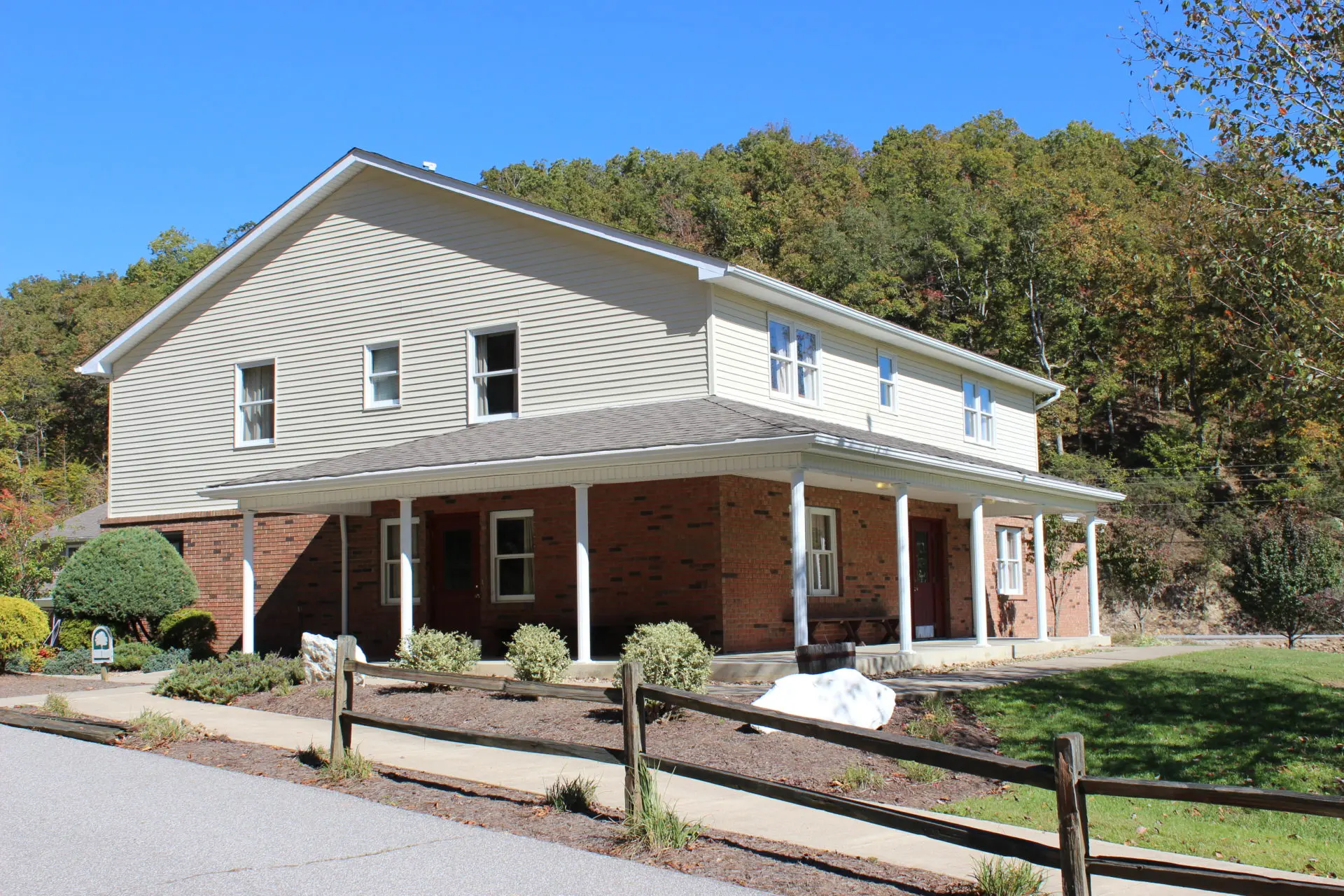 Two-story brick and siding building with a front porch and white columns, surrounded by trees and a wooden fence