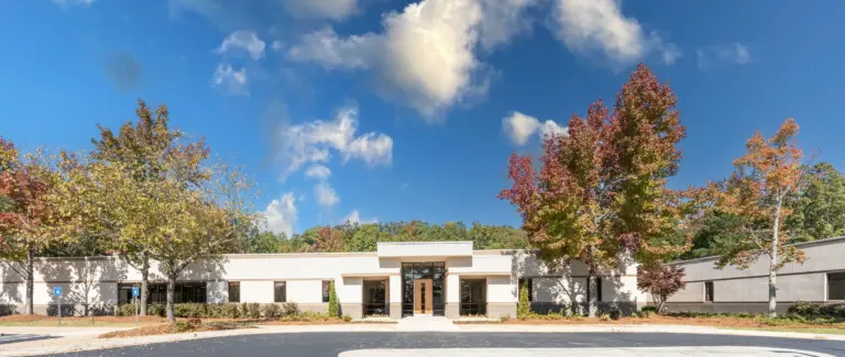 Single-story white rehab building with glass entrance and autumn trees under blue sky