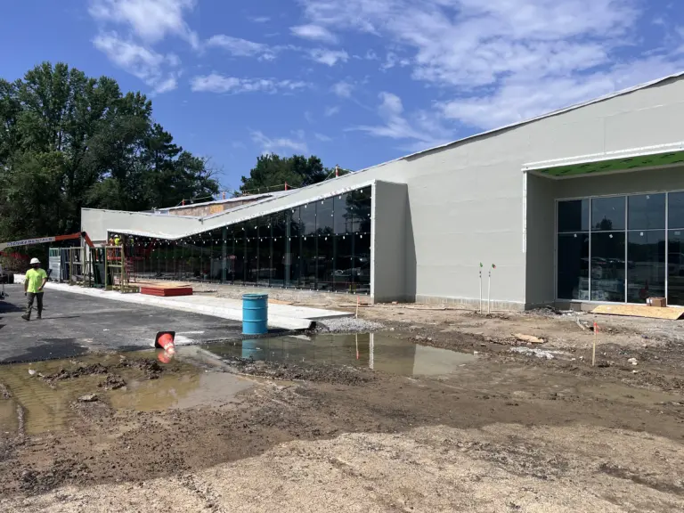 Newly built gray campus building with glass front under construction, with blue sky and trees behind