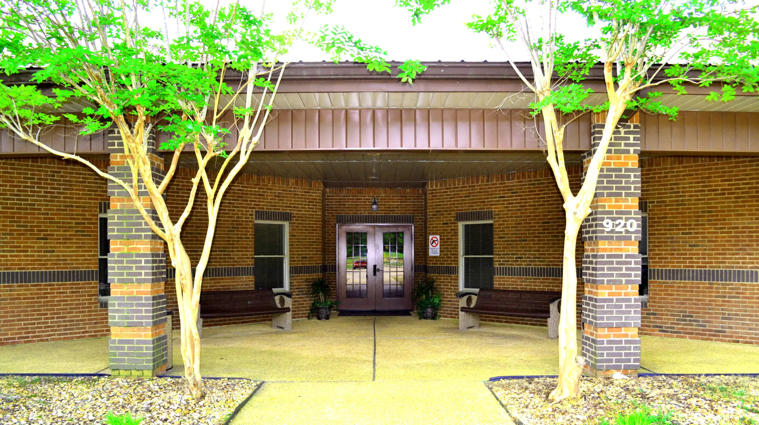 Brick building entrance with double glass doors, benches, and trees, with address number 920 on a pillar